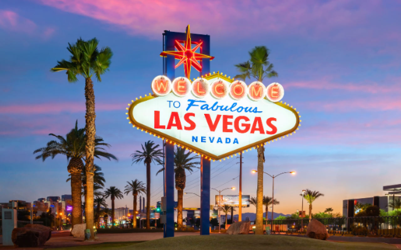 Il celebre cartello illuminato "Welcome to Fabulous Las Vegas Nevada" circondato da palme con il cielo rosa e viola del tramonto e le luci della Strip sullo sfondo