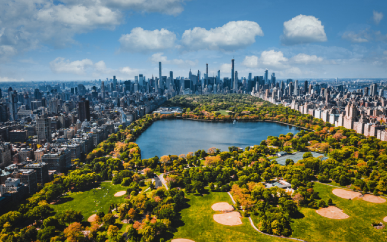 Vista aerea di Central Park in autunno con il laghetto circondato da alberi verdi e dorati e lo skyline dei grattacieli di Midtown Manhattan che si erge sullo sfondo sotto un cielo azzurro