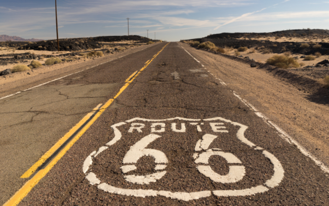 Il logo della Route 66 dipinto in bianco sull'asfalto di una strada rettilinea che si perde all'orizzonte tra il deserto e i pali del telegrafo, simbolo del viaggio on the road americano