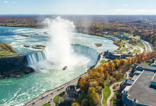 Vista aerea delle Cascate del Niagara in autunno: l'Horseshoe Falls a ferro di cavallo con la nuvola di spruzzi, la barca Maid of the Mist e i boschi colorati sulle due sponde