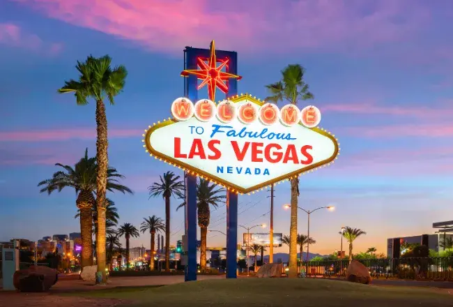 Il celebre cartello illuminato "Welcome to Fabulous Las Vegas Nevada" circondato da palme con il cielo rosa e viola del tramonto e le luci della Strip sullo sfondo