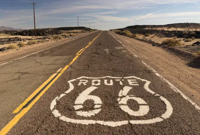 Il logo della Route 66 dipinto in bianco sull'asfalto di una strada rettilinea che si perde all'orizzonte tra il deserto e i pali del telegrafo, simbolo del viaggio on the road americano